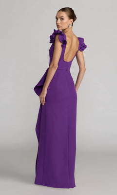 A woman stands in a studio setting, looking over her shoulder, wearing the Mac Duggal 27740 long formal dress—a floor-length, sleeveless purple gown featuring ruffled shoulders, a square neckline, and a low back.