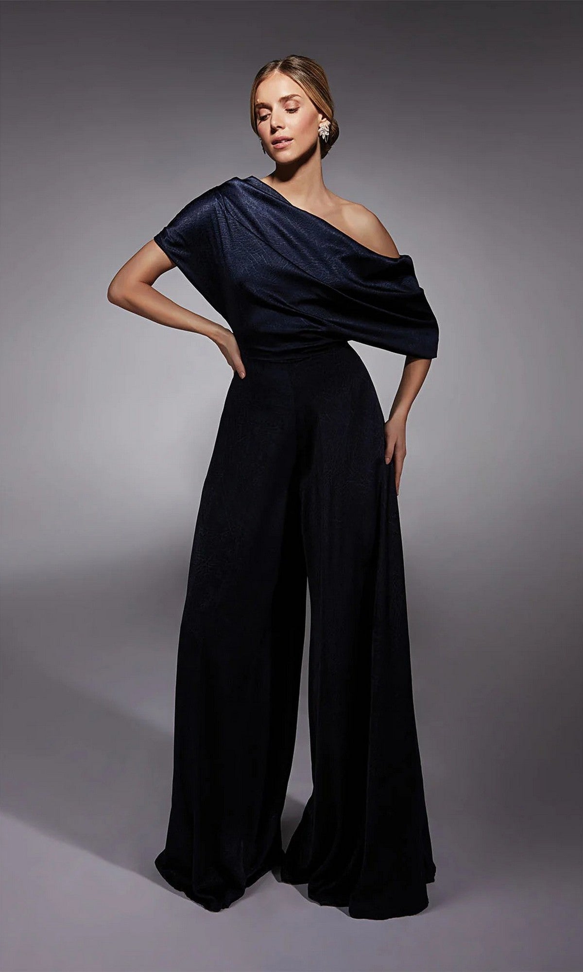 A woman models the Alyce 27743 long formal jumpsuit with off-the-shoulder style and wide-leg pants, ideal for a modern mother-of-the-bride look, against a simple grey background.