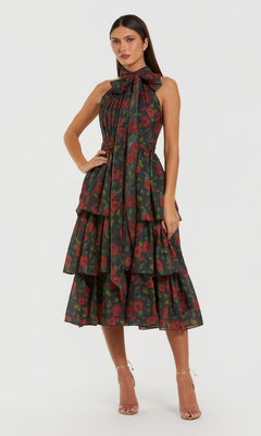 A woman poses in the Midi Wedding Guest Dress: Mac Duggal 27747, a black multi tea-length dress with a floral pattern and bow detail at the neck, paired with strappy high-heeled sandals.