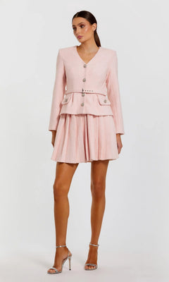 A woman stands wearing the Mac Duggal 27752 Short Boucle Day Dress, featuring a light pink textured design with embellished buttons, paired with silver high heels against a plain white background.