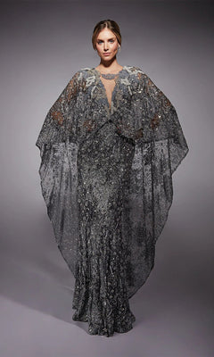 A person poses against a plain gray background wearing the Alyce 27752 long formal dress in charcoal, featuring floor-length glitter tulle and a sheer, wing-like cape.