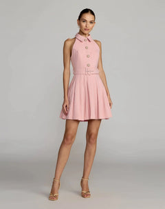 A woman wears the Short Party Dress: Mac Duggal 27766 in blush—a sleeveless collared design with large buttons and a matching belted waist—paired with heels against a plain gray background, embodying a chic, modern silhouette.