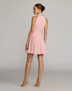 A woman stands facing away, wearing the Mac Duggal 27766 Short Party Dress—a sleeveless, collared light pink dress with a flared skirt—paired with beige high-heeled sandals against a plain, light background.