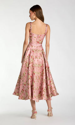 A woman stands facing away, wearing the Mac Duggal 27767 Midi Party Dress—a sleeveless, pink brocade party dress with a floral pattern, fitted bodice, and flared midi-length skirt—paired with light-colored high-heeled sandals.