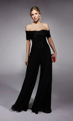 A woman in the Alyce 27768 Long Formal Jumpsuit in black with wide legs, paired with a red clutch and pearl earrings, stands against a plain gray background.