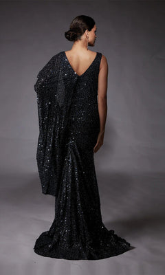 A woman faces away, wearing the shimmering Alyce 27772 long formal dress—a black sequin-beaded gown with a draped design and V-shaped back—standing against a plain gray background.