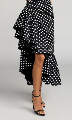 A person wearing the Mac Duggal 27780 Midi Party Dress—a black cocktail dress with a high-low ruffled skirt adorned with white polka dots—paired with black high-heeled sandals, photographed against a plain background.