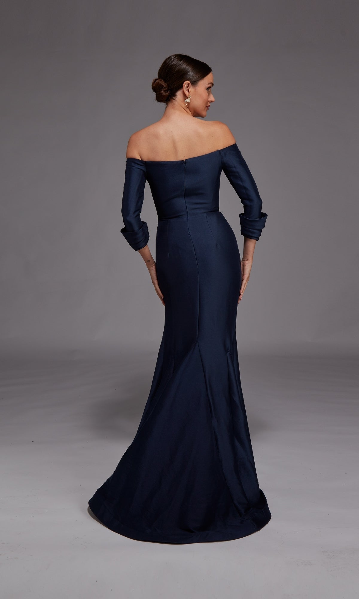 A woman in the Alyce 27782 Long Formal Dress stands facing away with her hair in a low bun against a neutral gray background.