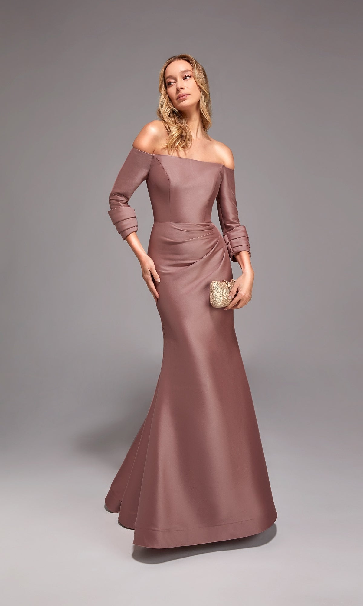A woman in the Alyce 27782 mauve off-shoulder mermaid dress with long sleeves holds a gold clutch while standing against a plain gray background.