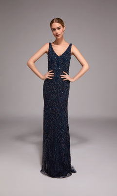 A woman poses in a studio wearing the Alyce 27786 long formal dress, a navy sleeveless beaded column gown with a V-neckline and floor-length design, her hands resting on her hips.
