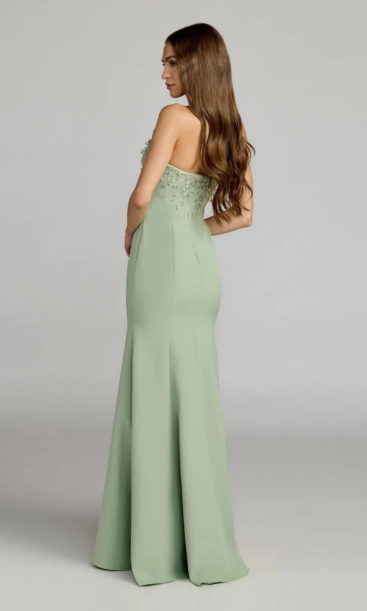 A woman wearing the Mac Duggal 27788 long formal dress—strapless and sage green with floral embroidery and delicate rhinestone embellishments on the bodice—poses in profile, showing off its elegant mermaid silhouette against a simple gray background.
