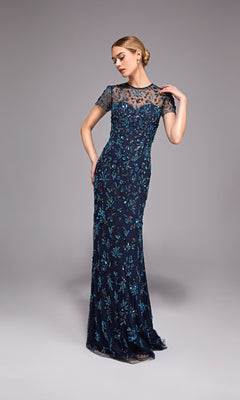 A woman in the Alyce 27789 long formal dress—navy blue, floor-length, beaded with short sleeves and blue floral embroidery—stands against a plain grey background.