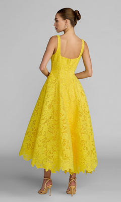 A woman is seen from the back wearing the Tea-Length Party Dress: Mac Duggal 27801—a bright yellow sleeveless lace dress with a flared mid-calf skirt—paired with heeled sandals, standing against a plain grey background.