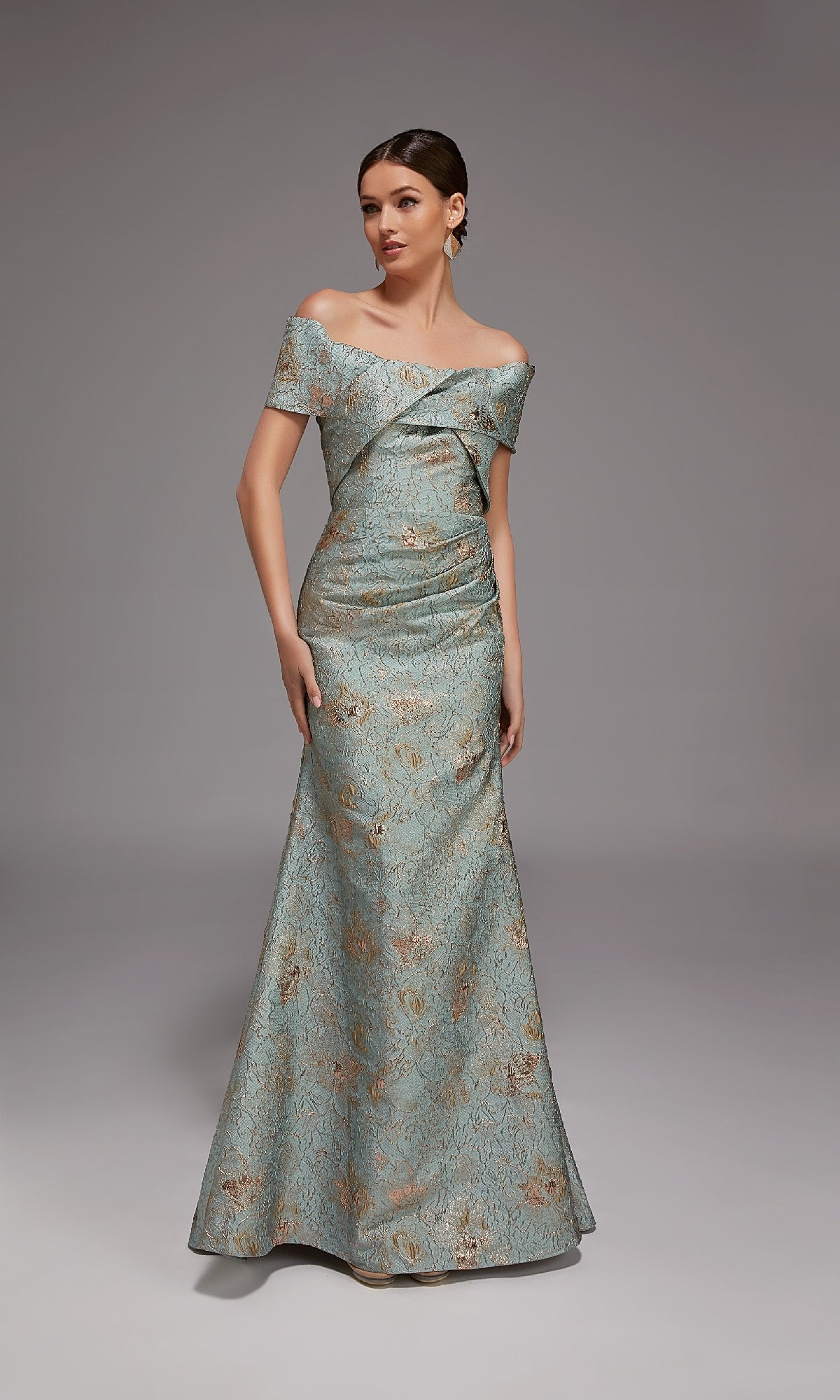 A woman wears the Alyce 27802 long formal dress, a floor-length, light blue off-the-shoulder gown with a subtle floral pattern, standing against a plain gray background.