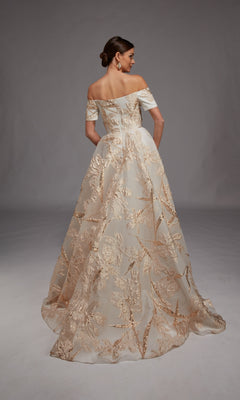 A woman faces away, wearing the Alyce 27804 long formal dress—an off-the-shoulder, floor-length gown featuring gold floral embroidery and metallic leaf motifs on a sheer overlay.