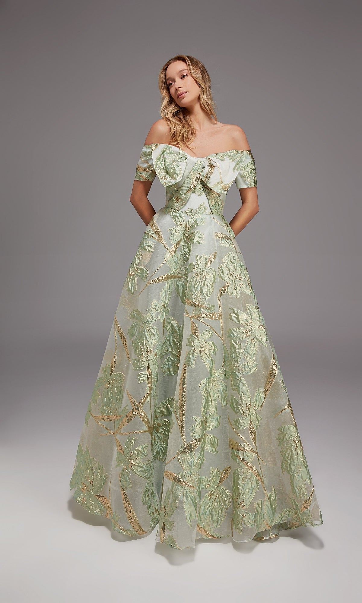 Wearing the Alyce 27804 long formal dress, a woman stands against a plain gray background. The gown features an off-the-shoulder design, floor-length silhouette, and green and gold metallic leaf motifs on sheer fabric.