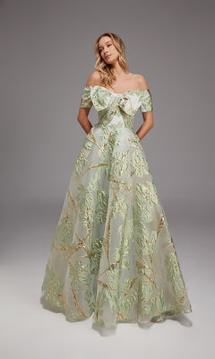 Wearing the Alyce 27804 long formal dress, a woman stands against a plain gray background. The gown features an off-the-shoulder design, floor-length silhouette, and green and gold metallic leaf motifs on sheer fabric.