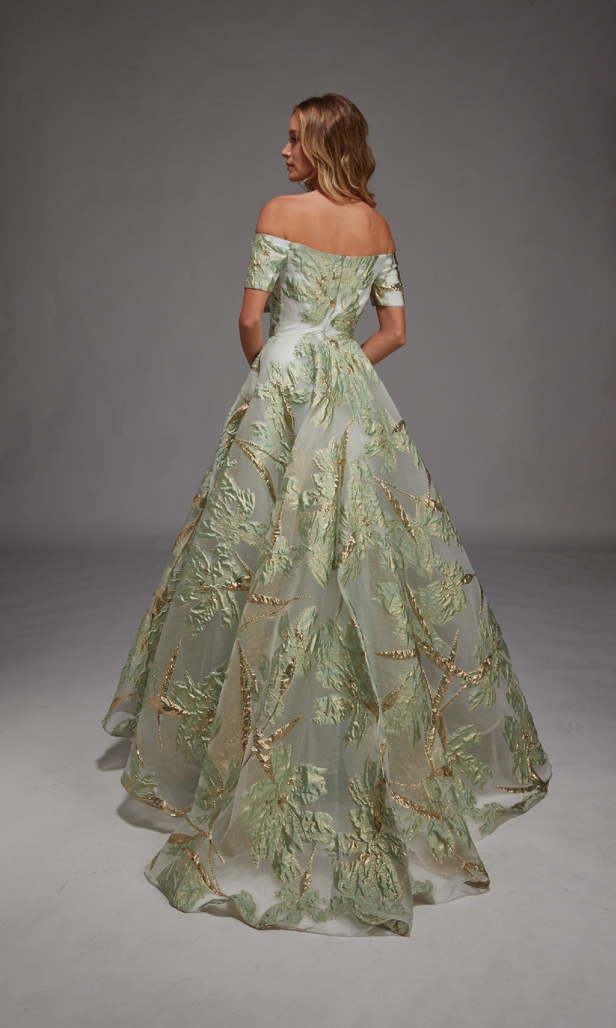 A woman faces away in a studio with a plain gray backdrop, wearing the Alyce 27804 long formal dress—an off-the-shoulder ball gown accented by green and gold metallic leaf designs.