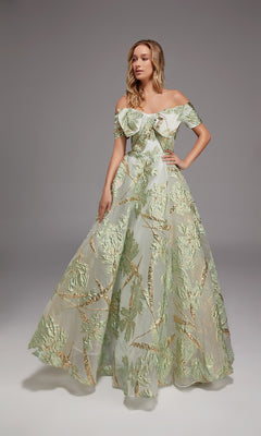 In a studio, a woman wears the Alyce 27804 long formal dress in Sea Glass/Gold, featuring an off-the-shoulder design and floor-length silhouette adorned with metallic leaf motifs.