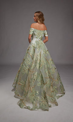 A woman in the Alyce 27804 long formal dress with off-shoulder design and green-gold metallic leaf motifs stands facing away against a plain gray background.
