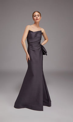 A woman poses in a studio wearing the Alyce 27806 Long Formal Dress, a strapless, floor-length charcoal gown with an elegant side bow detail.