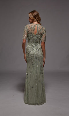 A woman faces away, wearing the Alyce 27809 long formal dress in light sage green, featuring sheer short sleeves and intricate beadwork—ideal for formal occasions or as a stunning mother-of-the-bride gown.