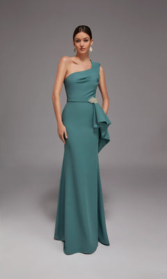 A woman stands in a studio wearing the Alyce 27812 long formal dress—a dark sage, one-shoulder, floor-length gown with a side drape and crystal beaded accent brooch at the waist.