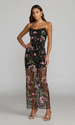 A woman wearing the Mac Duggal 27814 long formal dress in black multi—with pink floral embroidery, strapless neckline, sheer sections on the skirt—and gold heels, stands against a plain light gray background.