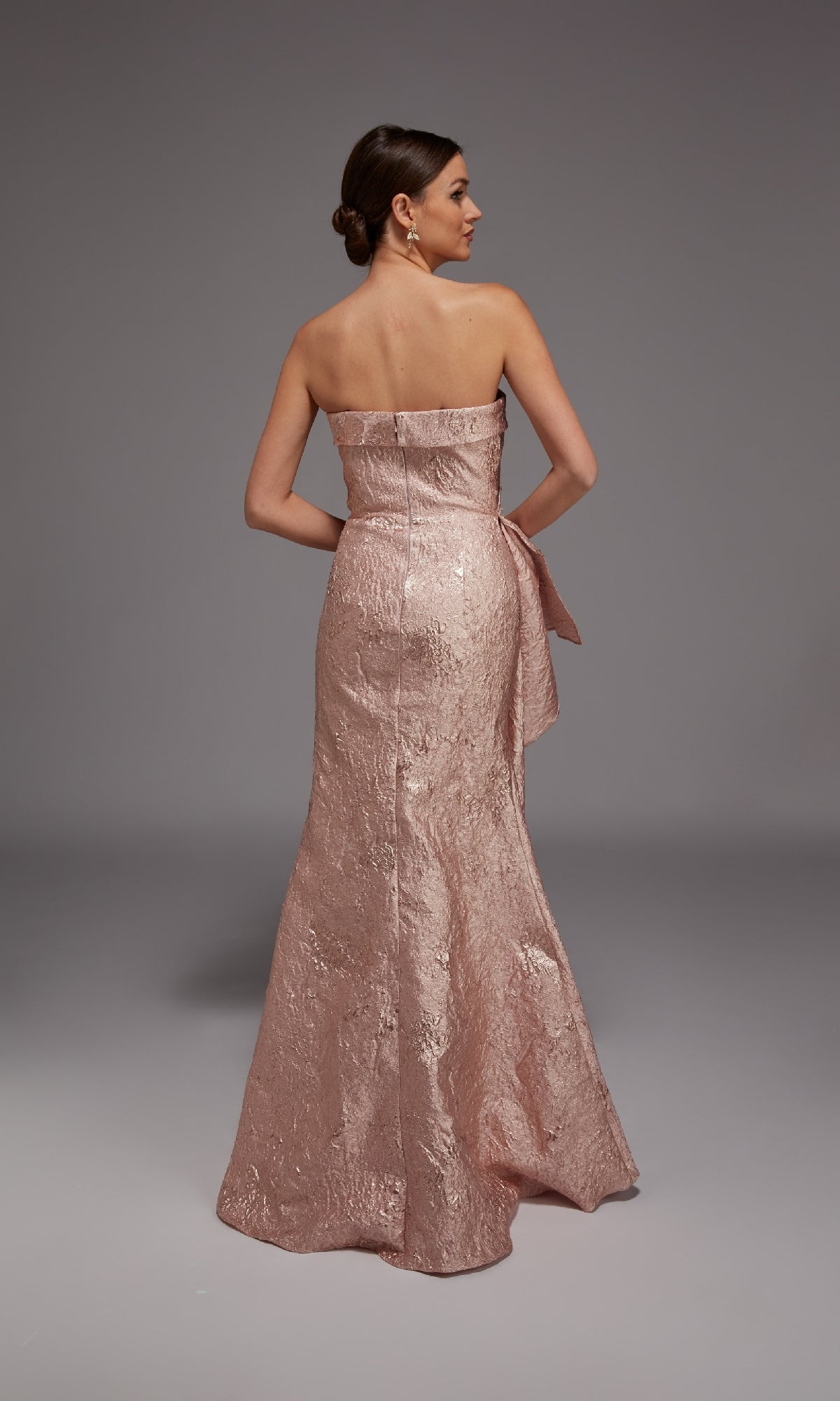 A woman faces away in the Alyce 27818 long formal dress—a strapless, light pink jacquard gown with a floor-length skirt and striking bow detail on the back, ideal for special occasions.