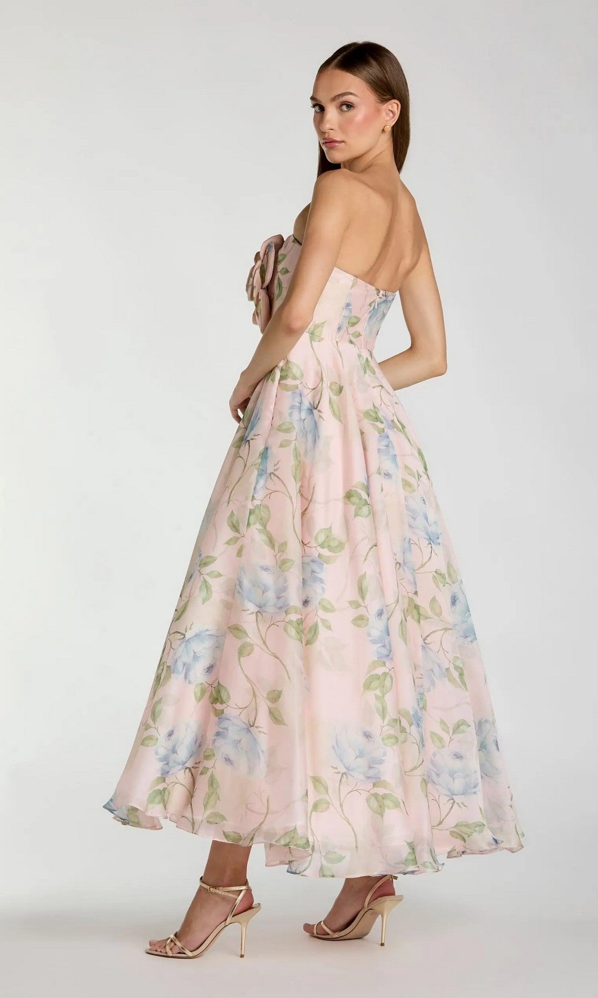 A woman stands sideways, looking over her shoulder in the Mac Duggal 27822 Midi Party Dress—a strapless, ankle-length pink floral organza dress with blue and green patterns and a delicate 3D flower bodice—paired with high-heeled sandals for an elegant look.