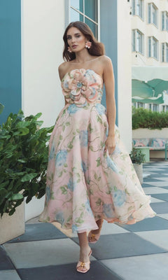 A woman in a strapless Powder Blue Multi Midi Party Dress: Mac Duggal 27822, featuring a 3D flower bodice and floral organza fabric, stands on a checkerboard patio near greenery and a building.