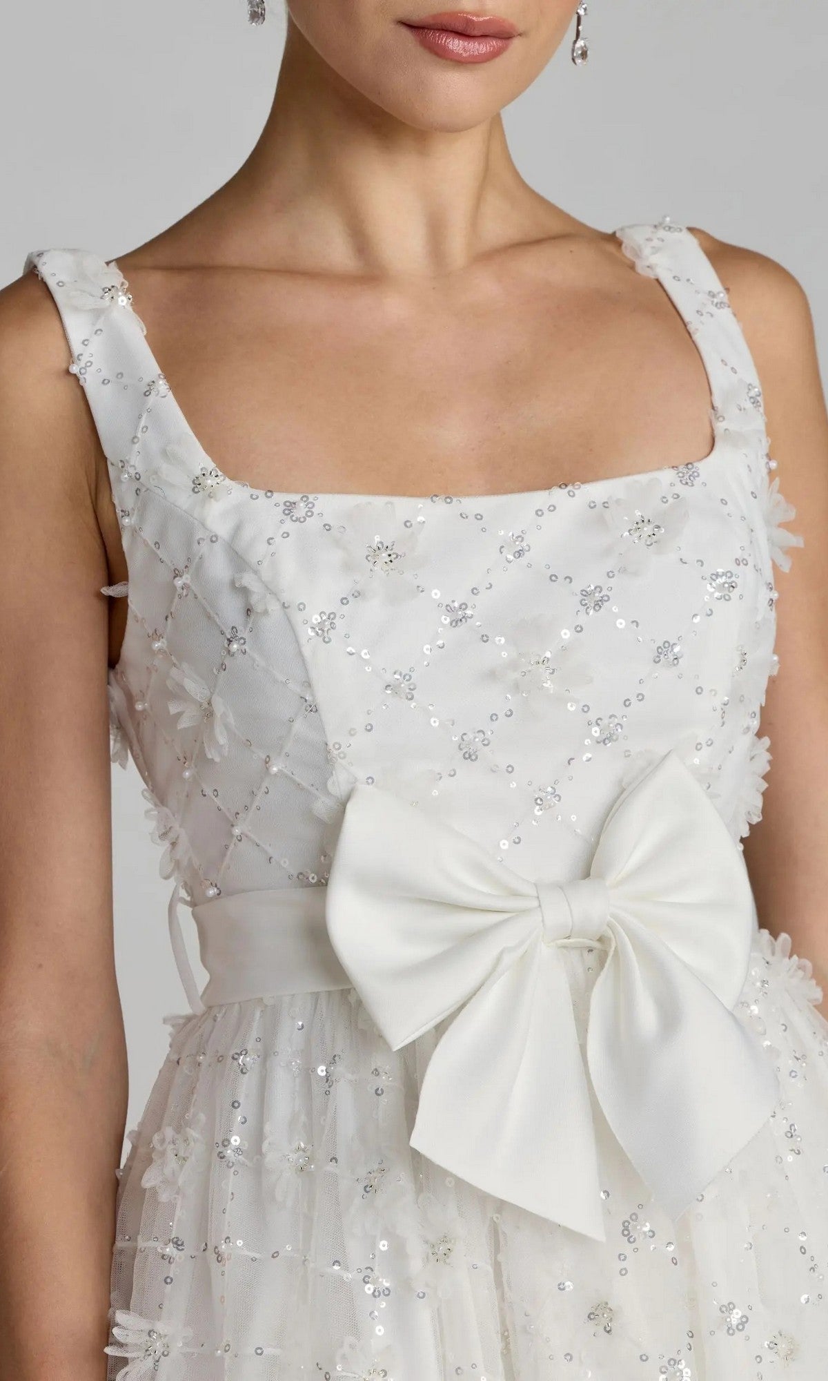 The Mac Duggal 27824 Midi Party Dress features white sequined embellishments, a square neckline, floral appliqués, and a large satin bow at the waist. Modeled with drop earrings, this tulle dress is perfect as a white midi party dress for any special occasion.