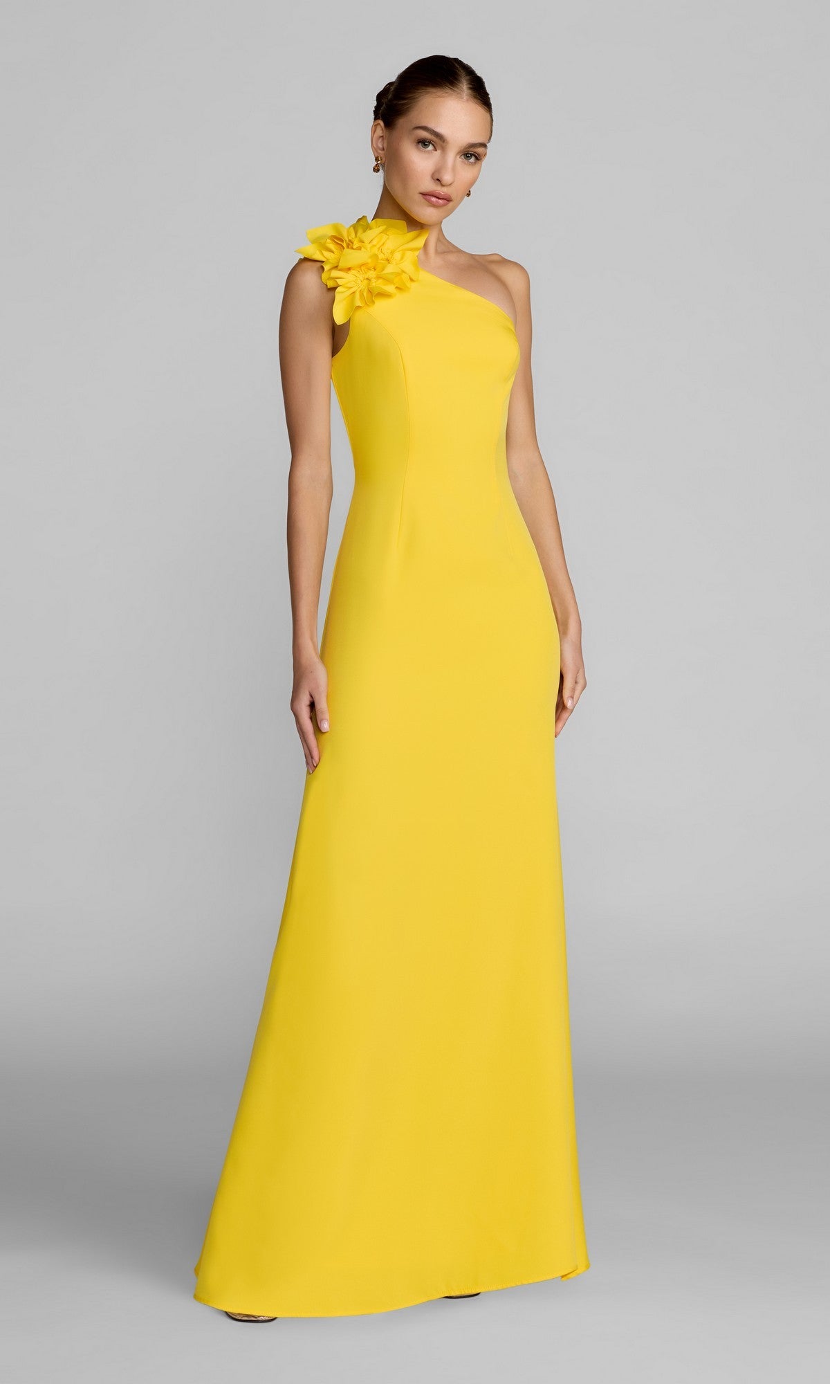 A woman stands against a plain gray background wearing the Mac Duggal 27825 long formal dress in Sunshine—a sleeveless, one-shoulder gown featuring an asymmetrical neckline and a large floral detail at the shoulder.