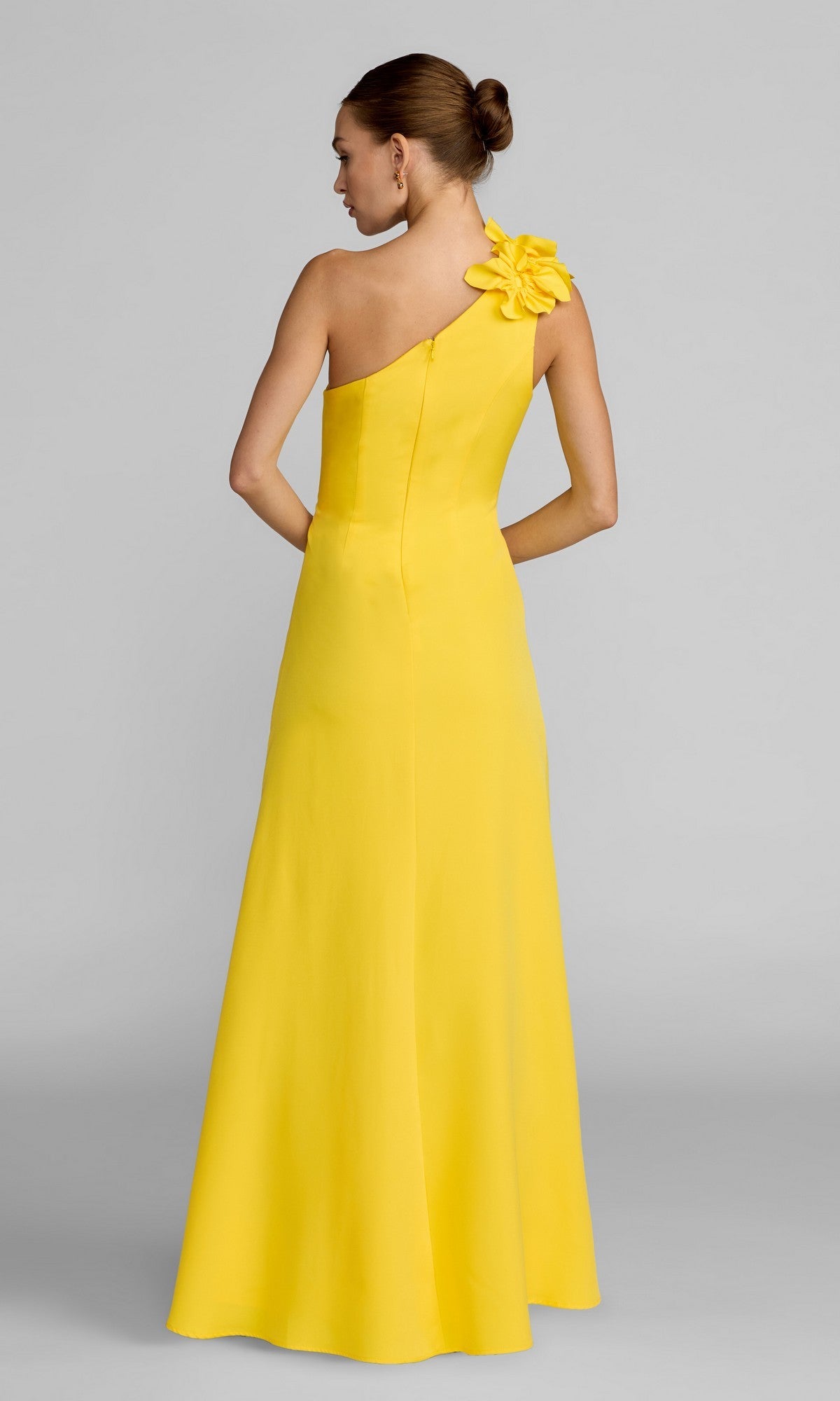 A woman stands with her back to the camera against a plain gray background, wearing the Mac Duggal 27825—a floor-length, yellow formal dress featuring a one-shoulder design and floral strap detail that channels timeless elegance.