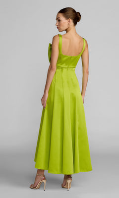 A woman stands in profile wearing the Mac Duggal 27826 midi party dress in apple-green satin, featuring a flared skirt and bow detail, paired with beige high heels.