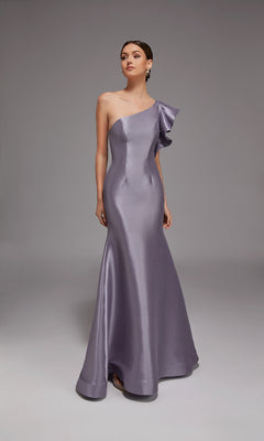 A woman stands against a plain gray backdrop wearing the Alyce 27826 long formal dress, a floor-length, one-shoulder metallic lavender gown with ruffled shoulder detail.