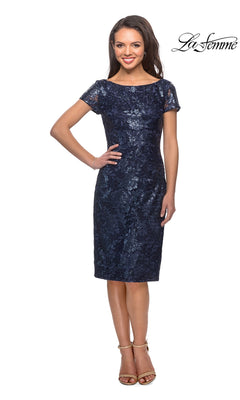 A woman wearing a navy blue Knee Length Wedding Guest Dress: La Femme 27828, featuring a knee-length, short sleeve lace design with a boat neckline.