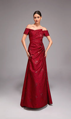 A woman poses in the Alyce 27828 Long Formal Dress—a floor-length, off-the-shoulder red jacquard gown with subtle floral patterns—against a plain gray background.