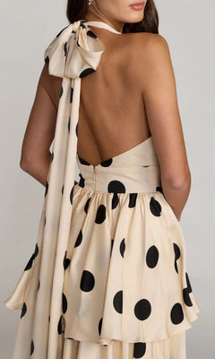 A woman wearing the Long Formal Dress: Mac Duggal 27833, an ivory midi dress featuring large black polka dots, an open back, a layered skirt, and a halter neckline accented with a bow tied from a long matching sash.