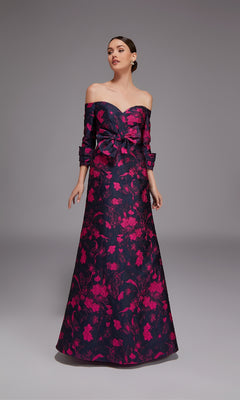 A woman poses against a dark background in the Alyce 27833, a long formal off-the-shoulder dress with an elegant A-line silhouette, bright pink floral pattern, floor-length hem, and a large bow at the waist.