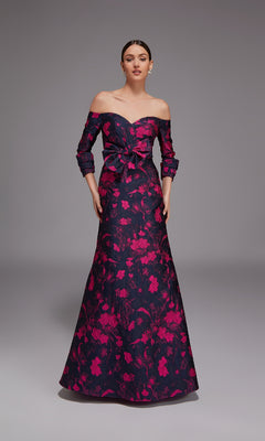 A woman wears the Alyce 27833 long formal dress, an off-the-shoulder jacquard gown with a dark base, bold pink floral print, fitted bodice, waist bow, A-line silhouette, and graceful floor-length hem.