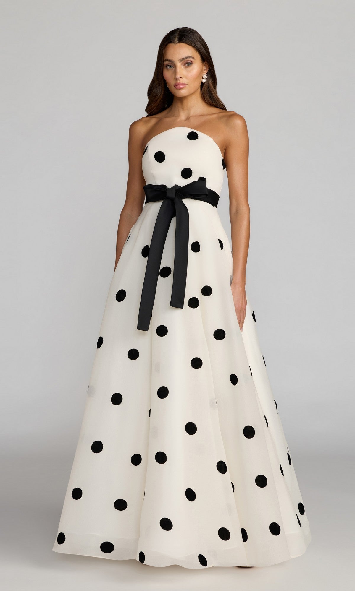 Wearing the Mac Duggal 27834 long formal dress in black ivory, a woman poses against a plain background; the strapless gown features bold black polka dots in organza and is finished with a black ribbon belt.