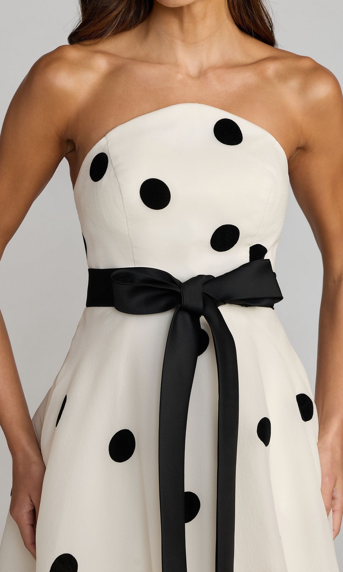 A woman wearing the Long Formal Dress: Mac Duggal 27834—a strapless white organza gown adorned with large black polka dots and finished with a black ribbon belt tied in a bow at the waist.