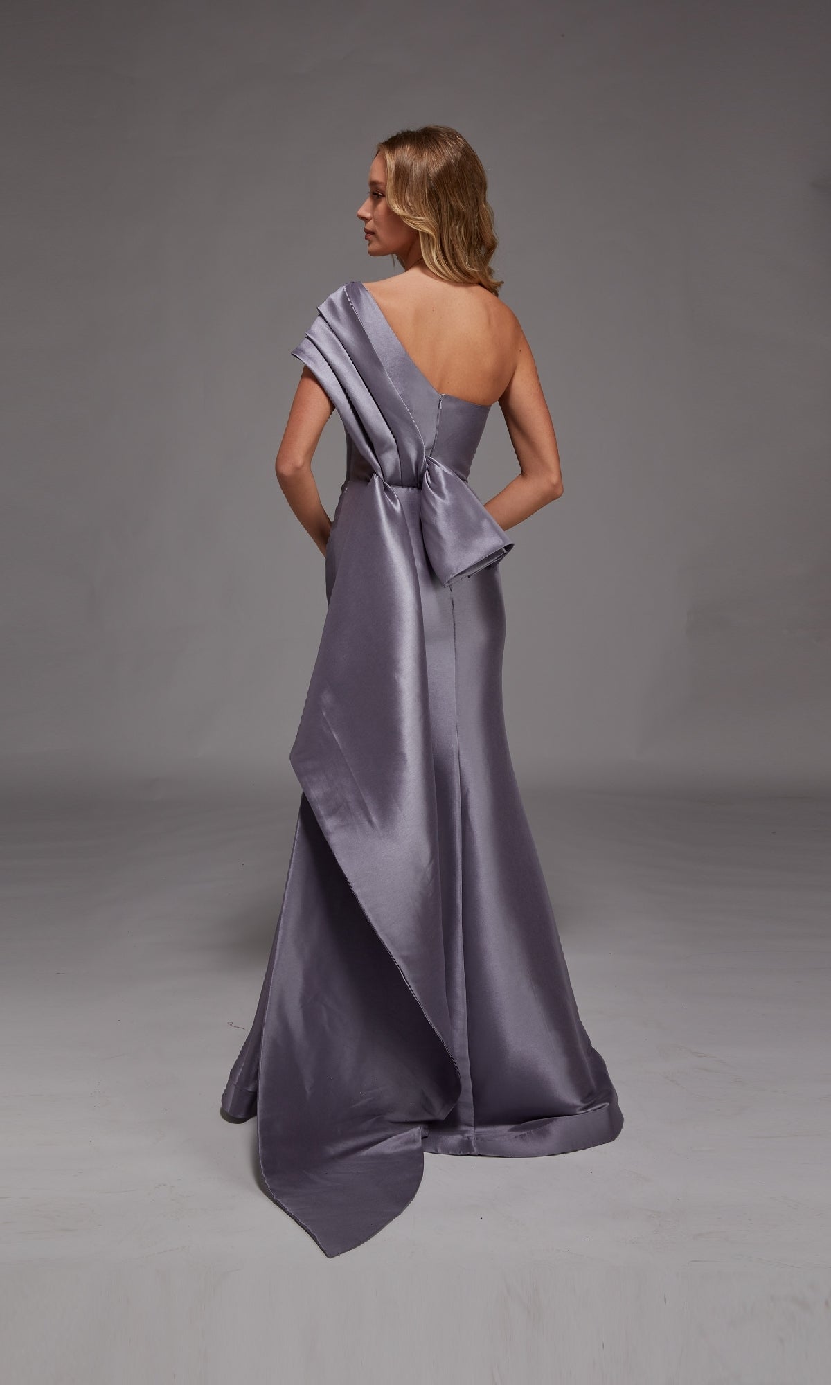 A woman stands on a plain gray background wearing the Alyce 27838, a floor-length, one-shoulder formal dress in silvery lavender with a large bow and draped fabric detail at the back.