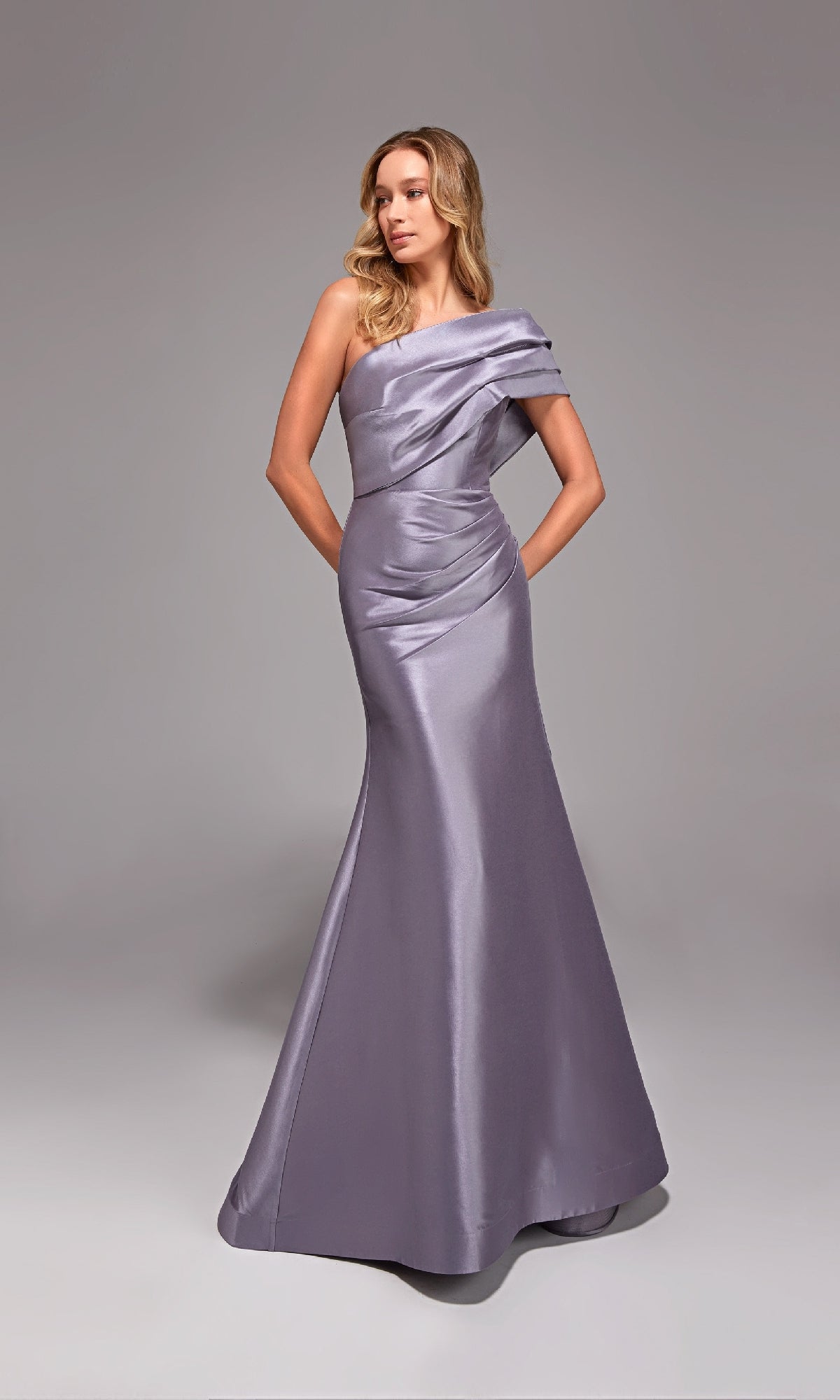 A woman stands against a plain gray background wearing the Alyce 27838 long formal dress, a shiny, floor-length, one-shoulder gown in lilac grey.