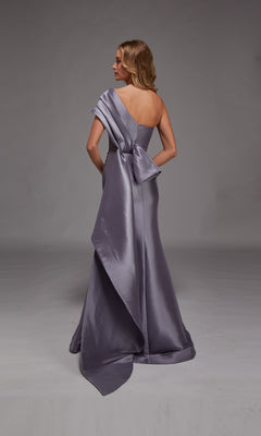 A woman faces away, wearing the Alyce 27838 long formal dress—a floor-length, one-shoulder silver satin gown with a large back bow detail—making it a stunning choice for any elegant event.