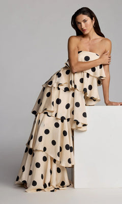 A woman wearing the Long Formal Dress: Mac Duggal 27845—a strapless, tiered cream gown adorned with large black polka dots—leans against a white rectangular block, striking a pose for a studio fashion photo.