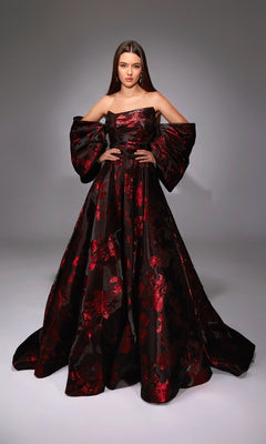 A woman poses in a studio wearing the Alyce 27862, a long formal off-the-shoulder floral jacquard gown with red patterns and dramatic puffed sleeves—an ideal choice for any formal ball.