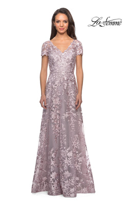 A woman wearing a floor-length, short-sleeve, antique blush La Femme 27870 mother of the bride dress with delicate floral lace patterns stands against a white background. This modest formal gown beautifully showcases intricate detailing, while the La Femme logo appears in the top right corner.