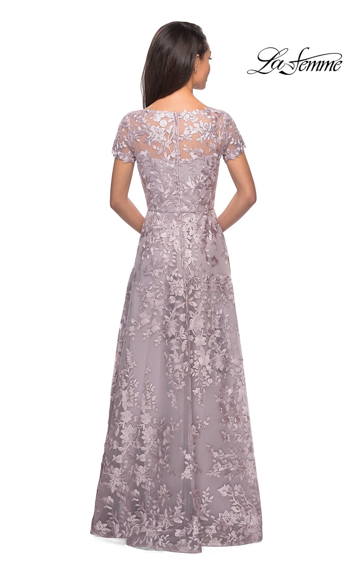 A woman stands with her back to the camera, wearing the La Femme 27870—a modest, floor-length mauve gown featuring short sleeves and a floral lace overlay. This elegant formal dress is perfect for mothers of the bride, with the “La Femme” logo displayed in the upper right corner.